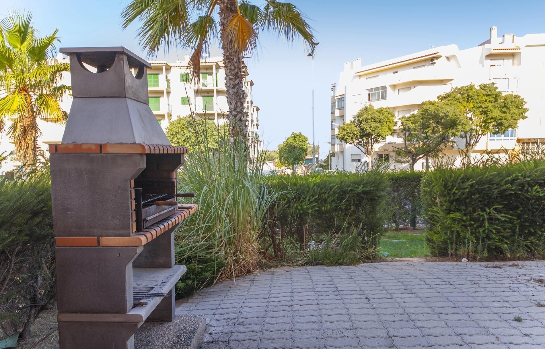 Un área de parrilla moderna al aire libre con una estructura de piedra, rodeada de follaje y edificios residenciales al fondo.