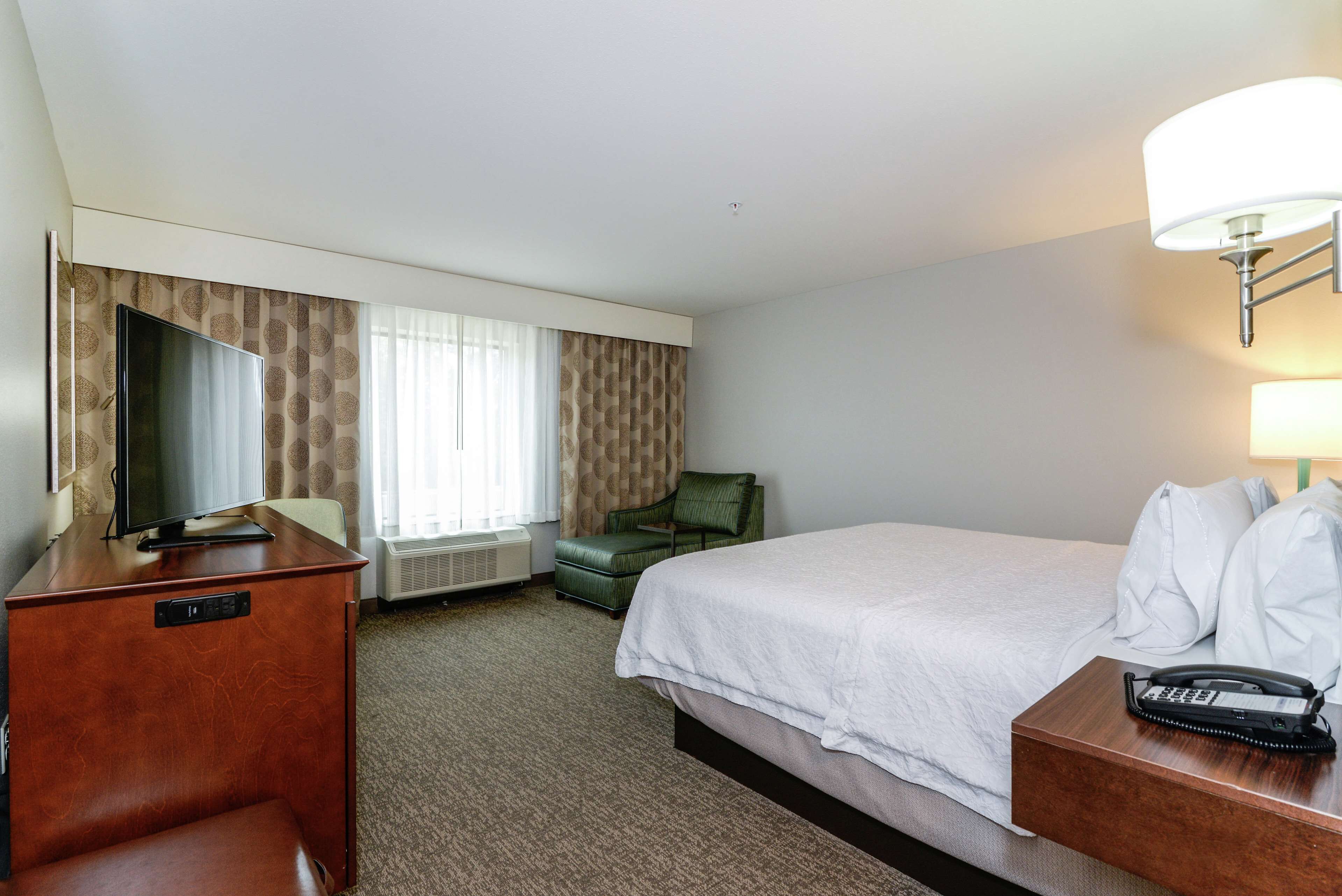 Habitación de hotel con cama, escritorio con televisor, silla, mesita de noche con teléfono y ventana con cortinas.