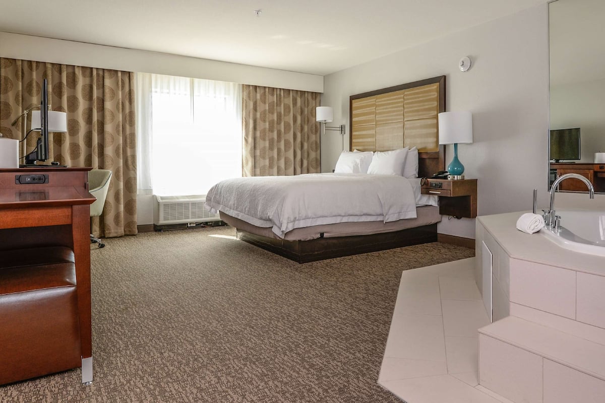 A hotel room with a bed, a desk, a chair, a desk lamp, a nightstand, a lamp, a bathroom with a bathtub, and a cityscape painting on the wall.