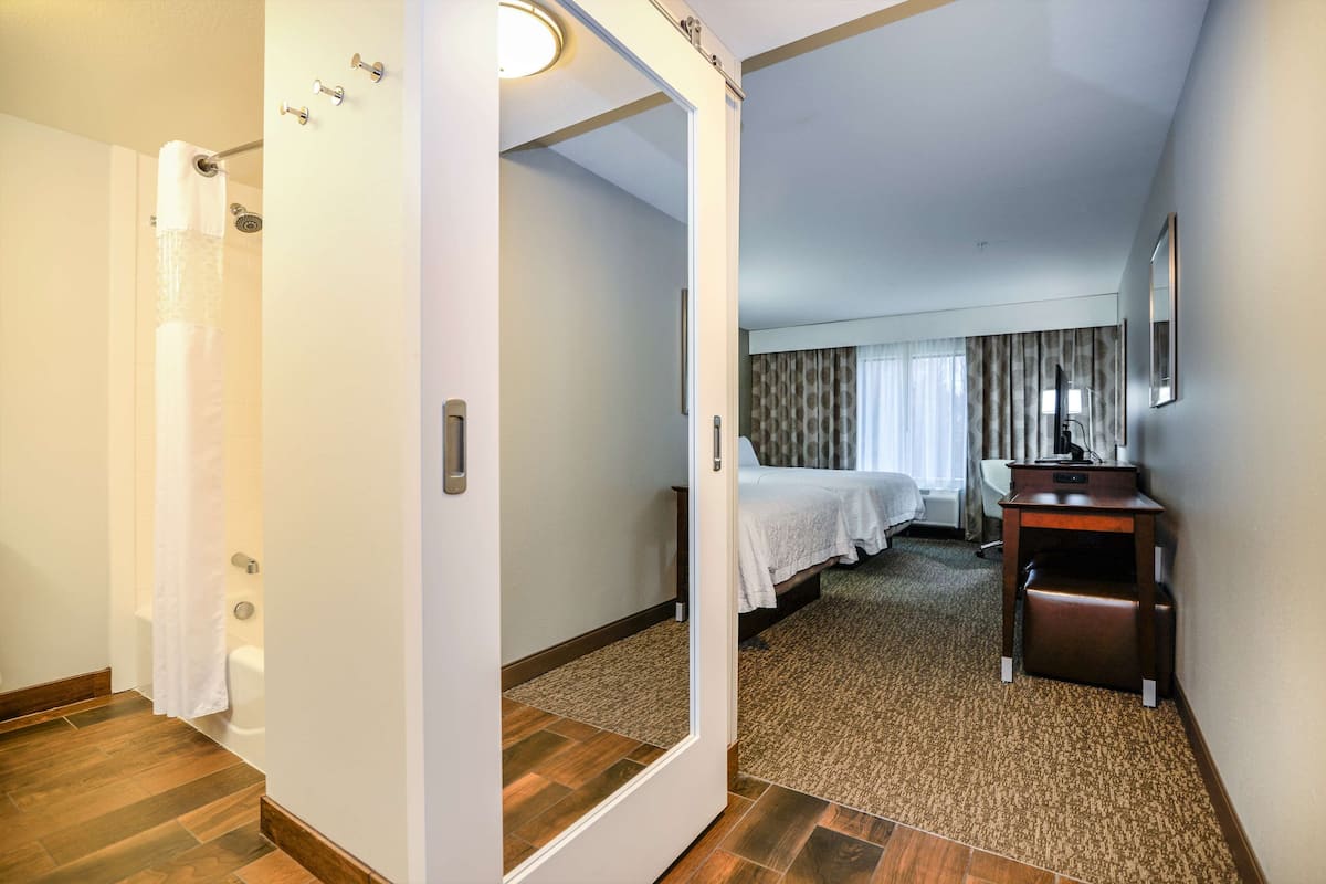A hotel room with a wooden floor, a bed with a white bedspread, a desk with a chair, and a shower area.