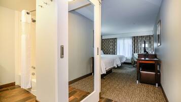 Une chambre d’hôtel avec un plancher en bois, un lit recouvert d’une courtepointe blanche, un bureau avec une chaise et un espace douche.