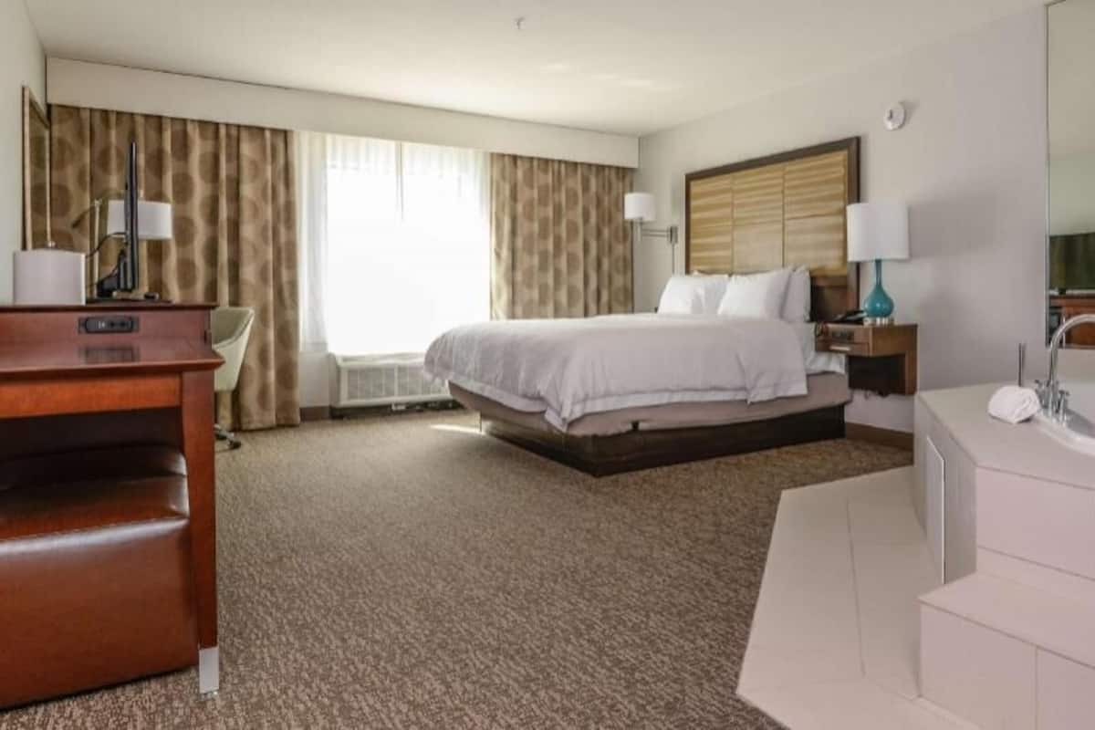 A hotel room with a bed, a desk, a chair, a large mirror, a bathtub, and a window with curtains.