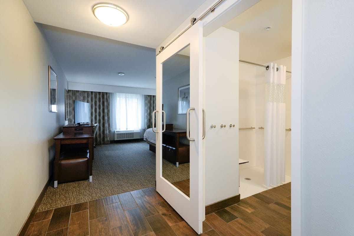 A hotel room with a wooden floor, a desk with a chair, a bed, a shower area, and a mirror.