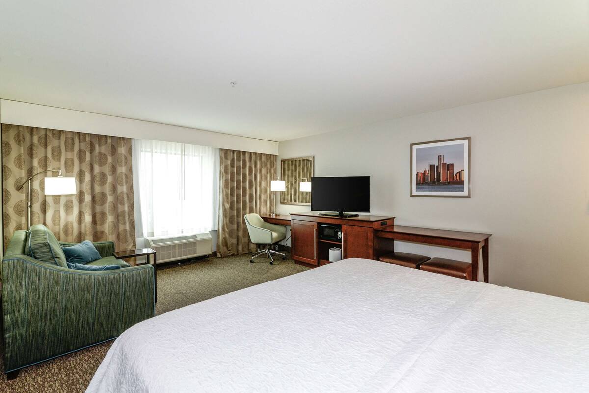 A hotel room with a large bed, a desk with a television, a chair, and a window with curtains.