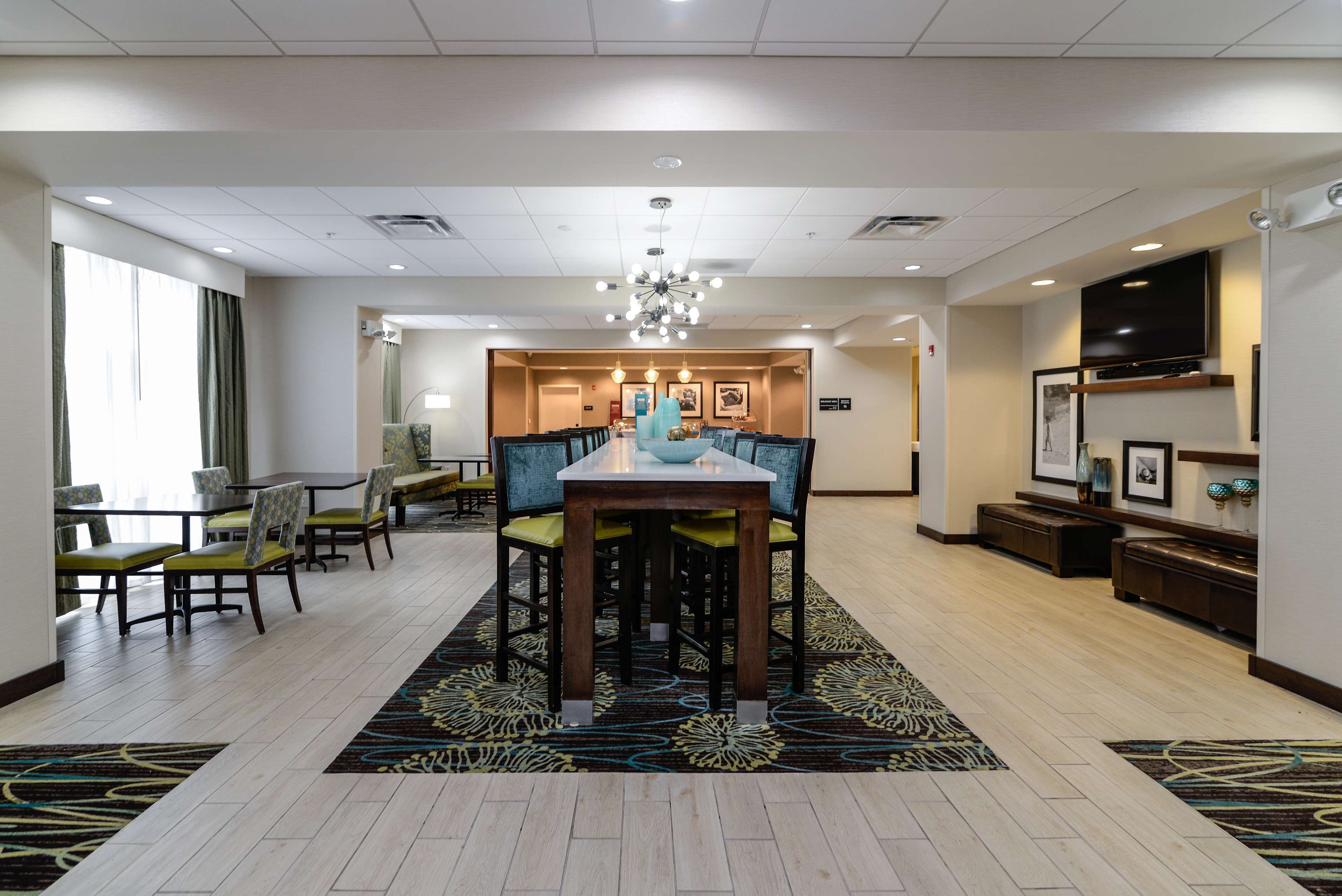 A hotel lobby with a dining area featuring a long table and chairs, a chandelier, and a television on the wall.