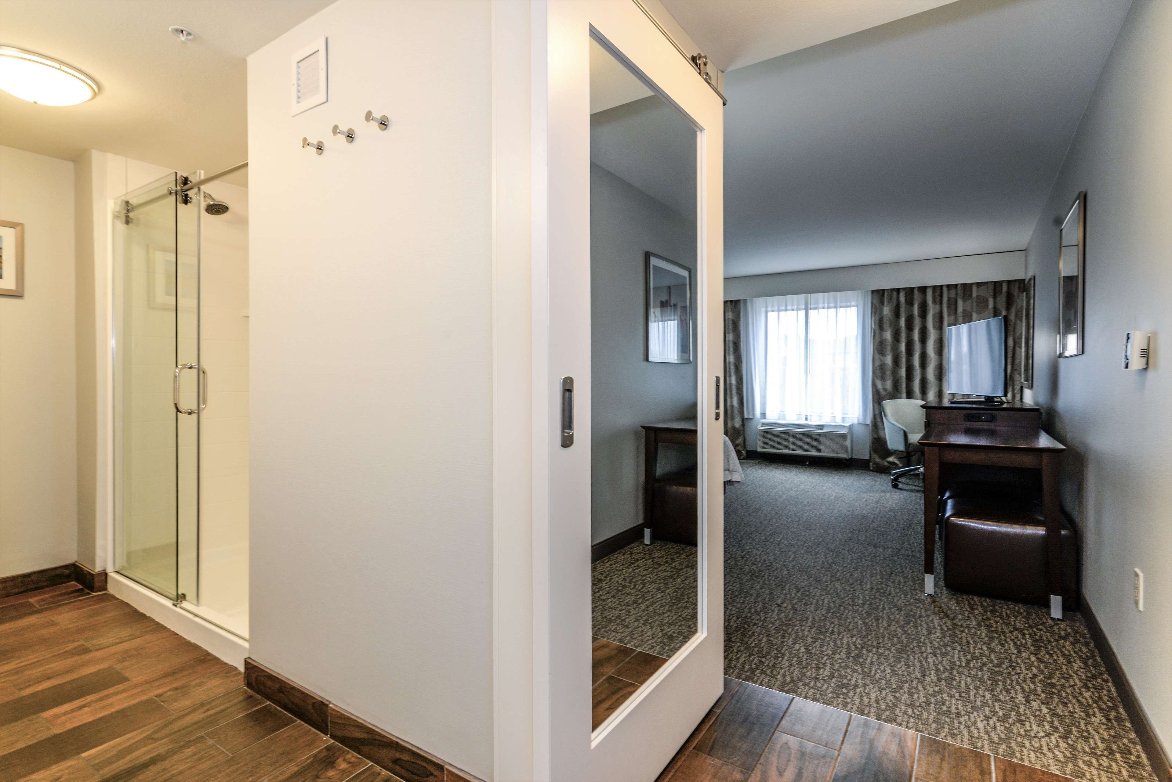 Habitación de hotel con ducha de vidrio, escritorio, silla y un espejo grande.
