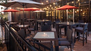 An outdoor seating area with wicker furniture and red umbrellas, featuring a modern building with large windows in the background.