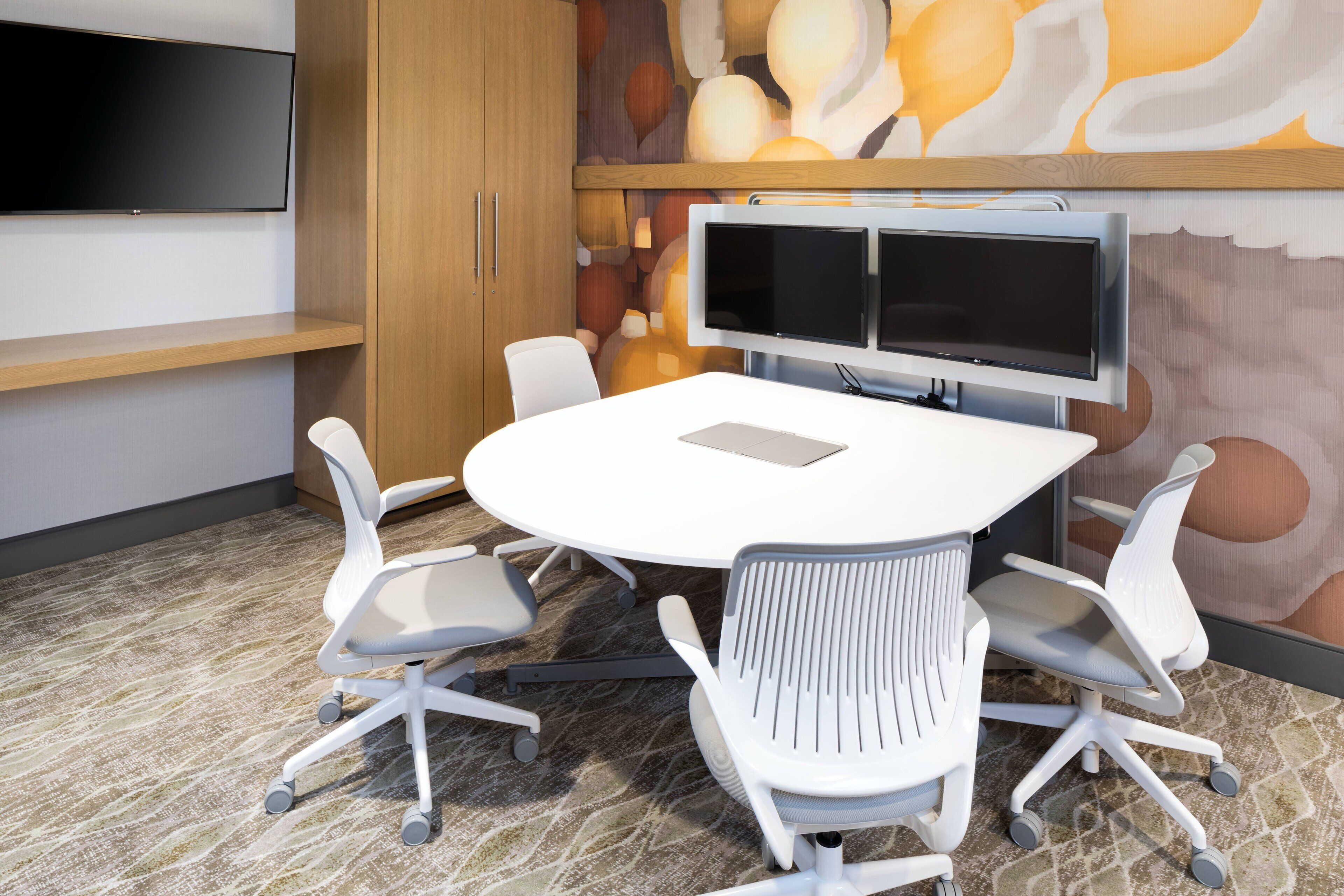 A conference room with a round table, four white chairs, a large flat-screen TV, and a wooden cabinet.