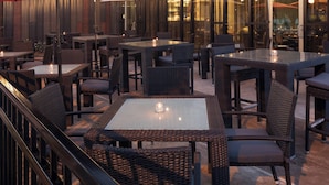 Un área de comedor al aire libre con mobiliario de mimbre y sombrillas rojas, con vista a un edificio moderno con el nombre