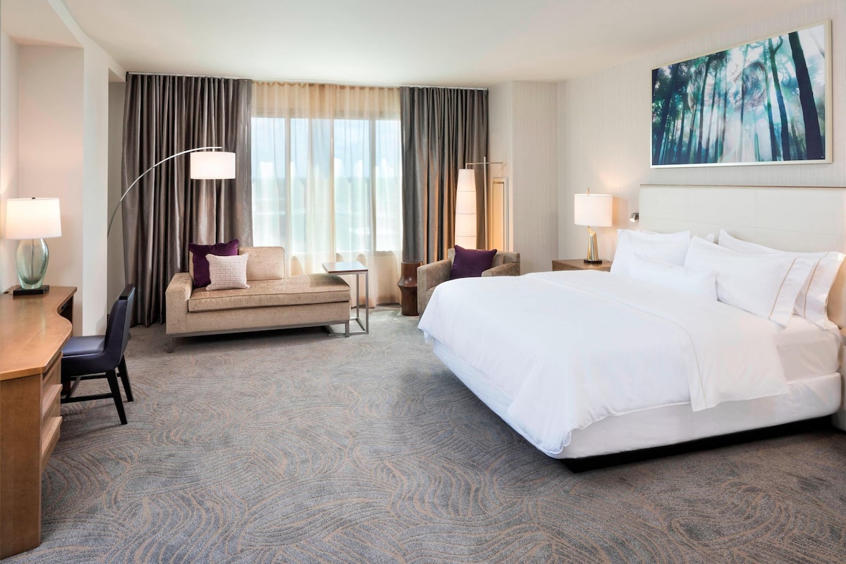 A hotel room with a large bed, a sofa, a desk with a chair, and a desk lamp. There is a window with curtains and a painting on the wall.