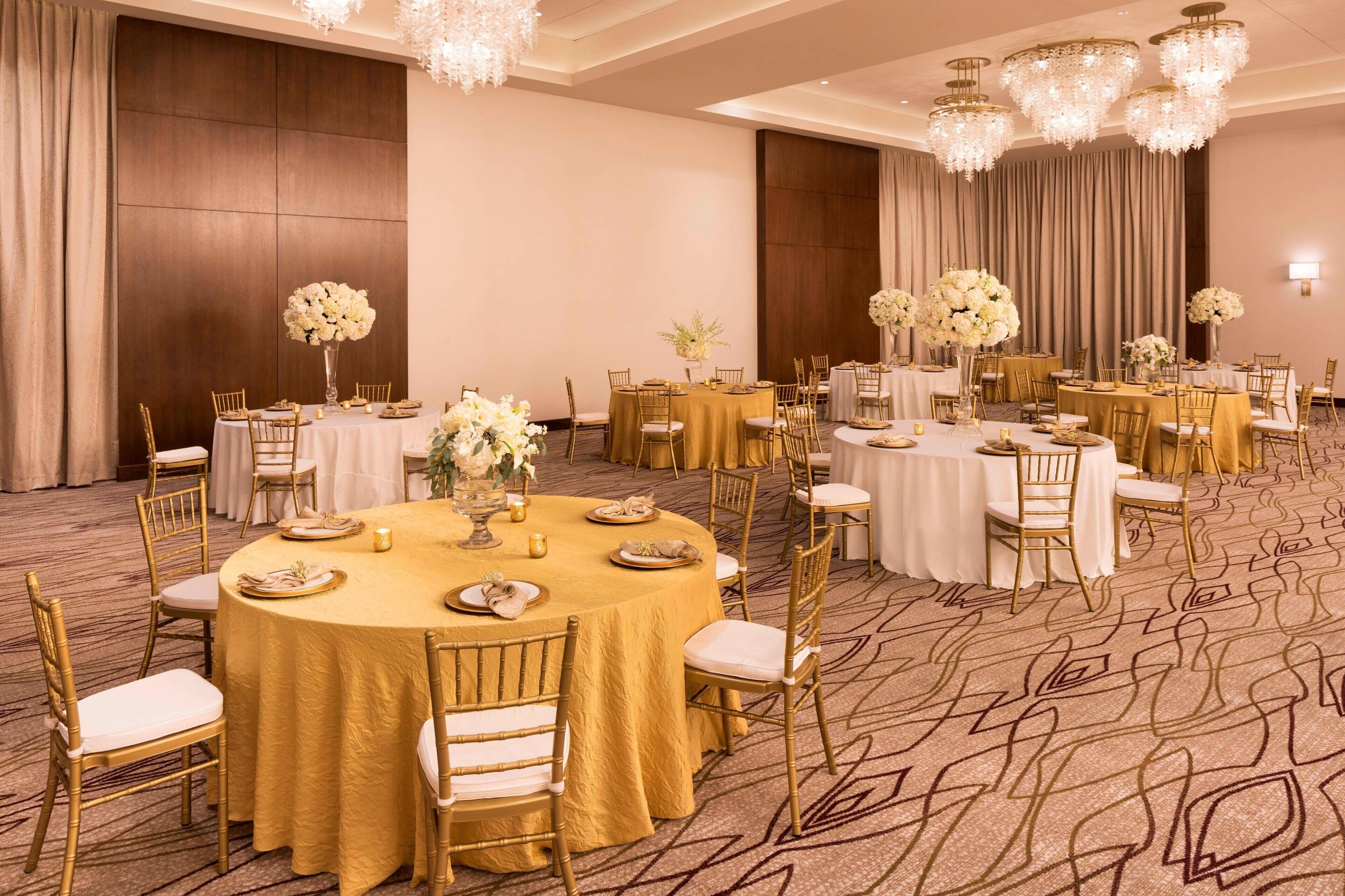 Une salle de banquet avec des tables rondes recouvertes de nappes dorées, des chaises blanches et des centres de table floraux.