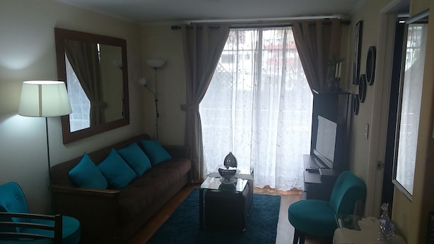A living room with a sofa, a coffee table, a lamp, a TV, and a window with curtains. - Departamento Belen Marcoleta (Santiago)