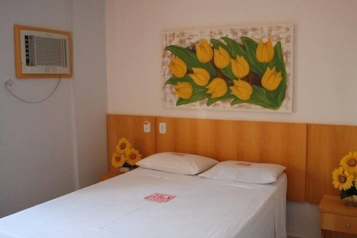 Ein Hotelzimmer mit Bett, Kopfteil, Wandklimaanlage und einem Sonnenblumenbild.