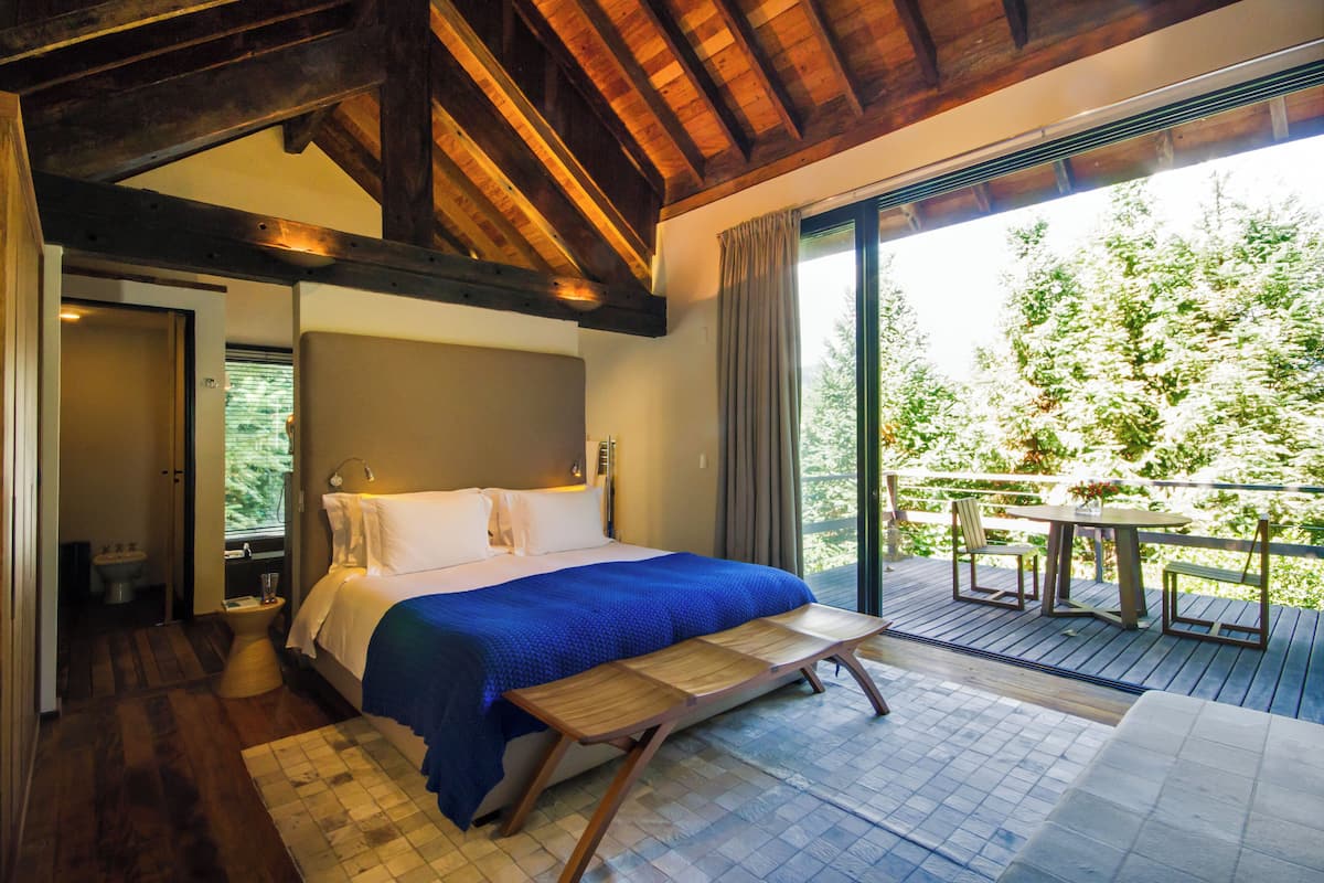 A bedroom with a large bed, wooden bench, and a view of a balcony with a table and chairs.