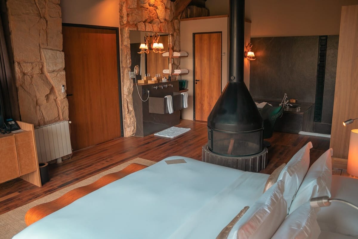 A bedroom with a stone wall, wooden floor, a bed with white linens, a fireplace, and a bathroom with a sink and towel rack.