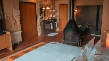 Un dormitorio con pared de piedra, suelo de madera, una cama con ropa blanca, chimenea y un baño con lavabo y toallero.