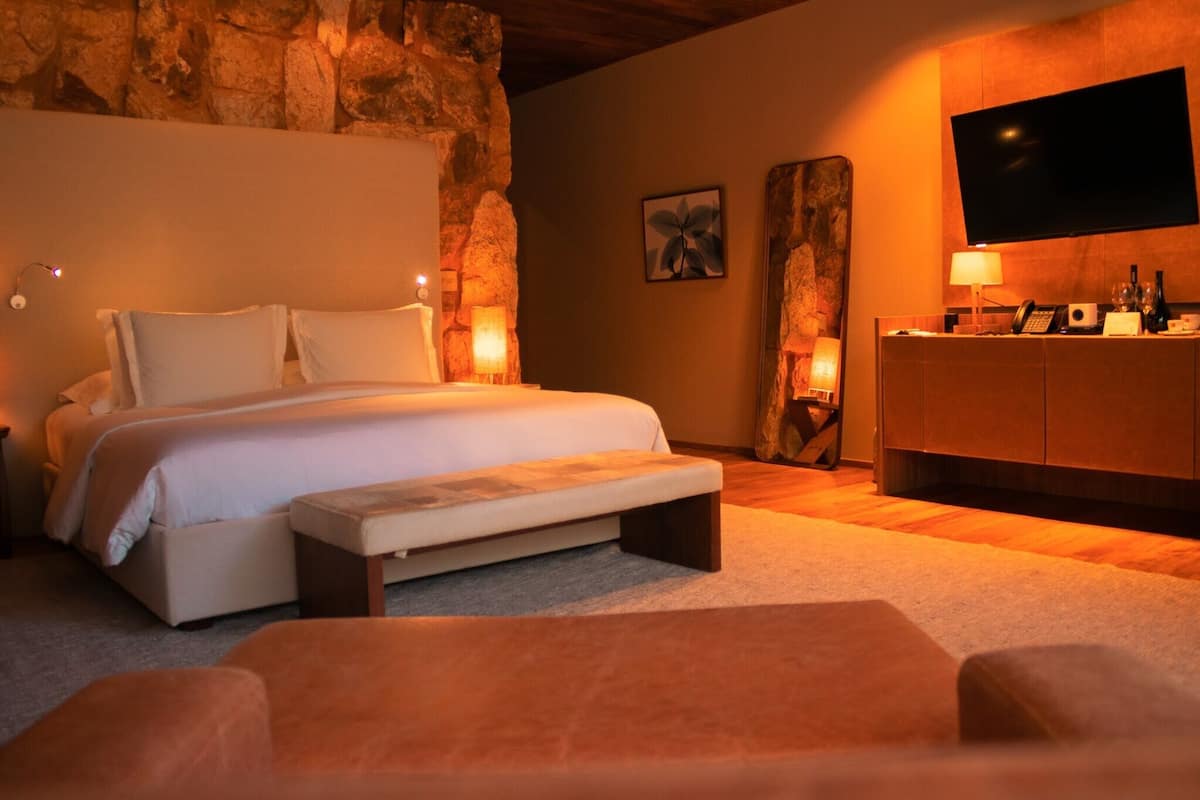 A bedroom with a large bed, bedside lamps, a flat-screen TV, a wooden dresser, and a stone wall.