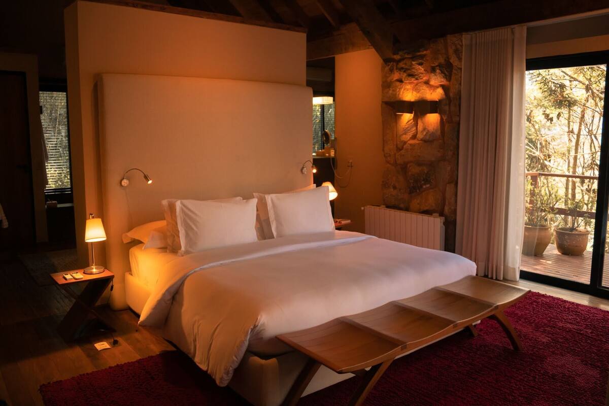 A bedroom with a large bed, a wooden bedside table, a nightstand, a red rug, and a view of trees through a window.