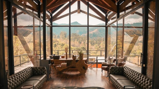 Ein geräumiges Zimmer mit großem Fenster und weitem Panoramablick auf die Berge. In der Mitte befinden sich zwei Ledersofas und ein Holztisch.