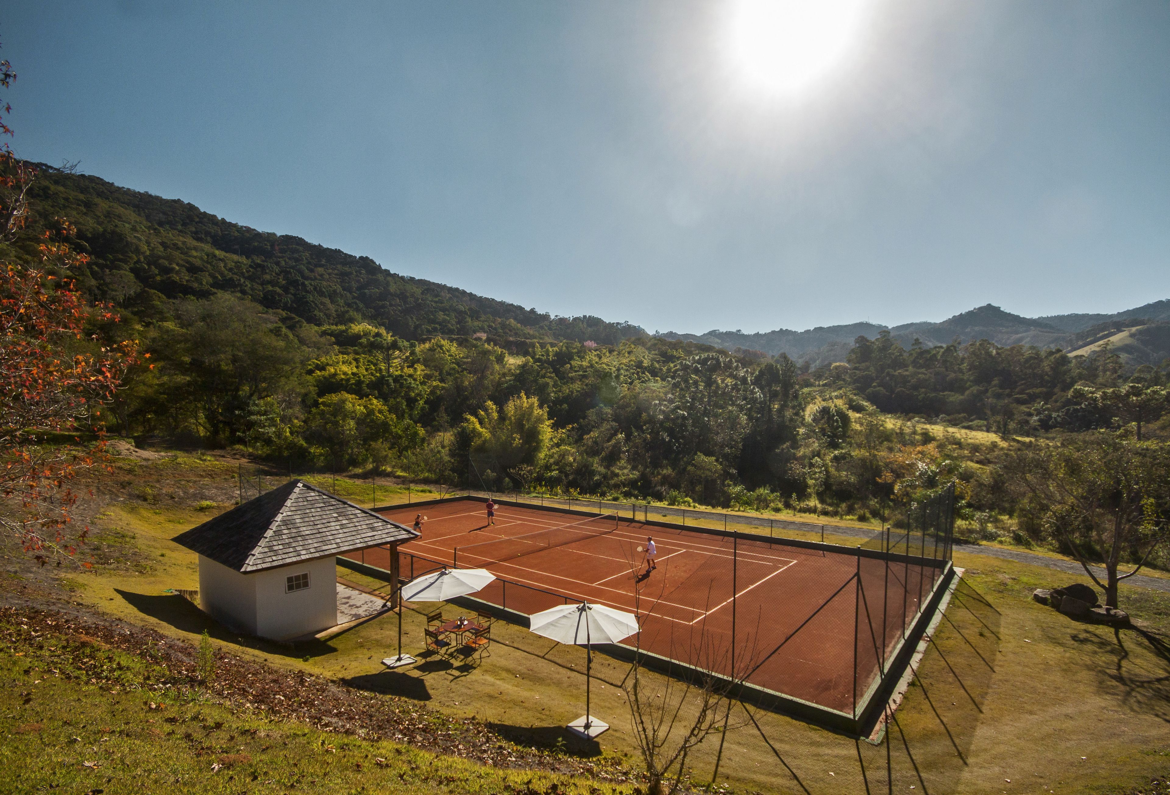 A tennis court with a small building and umbrellas, surrounded by hills and trees.