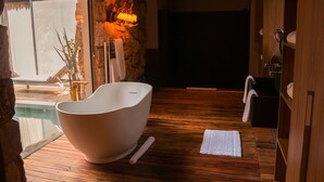Ein modernes Badezimmer mit freistehender Badewanne, Holzboden, einer Steinwand und einem Fenster mit Blick auf einen Poolbereich.