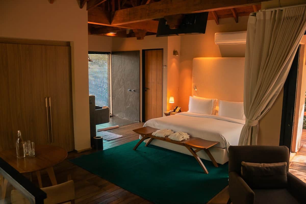 A hotel room with a bed, a bedside table with a lamp, a wooden bench, a chair, and a view of the outside through a door.