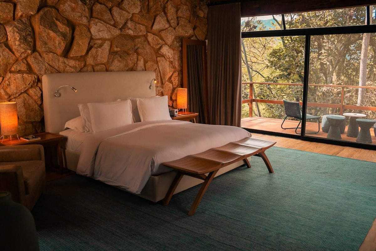 A bedroom with a stone wall, a large bed, two bedside lamps, a wooden bench, and a balcony with outdoor seating.