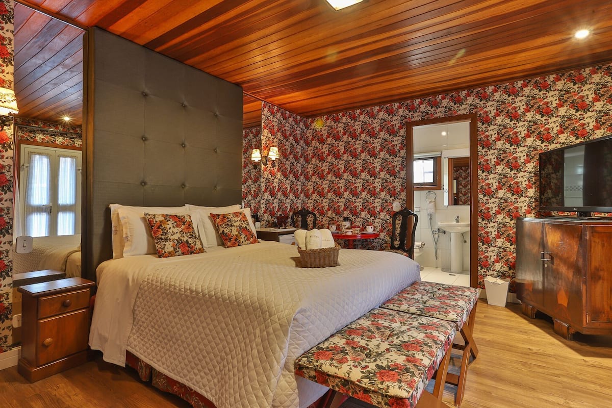 Une chambre à coucher avec un grand lit, une table de chevet, un banc, une armoire en bois, un téléviseur et une salle de bain visible à travers une porte ouverte.