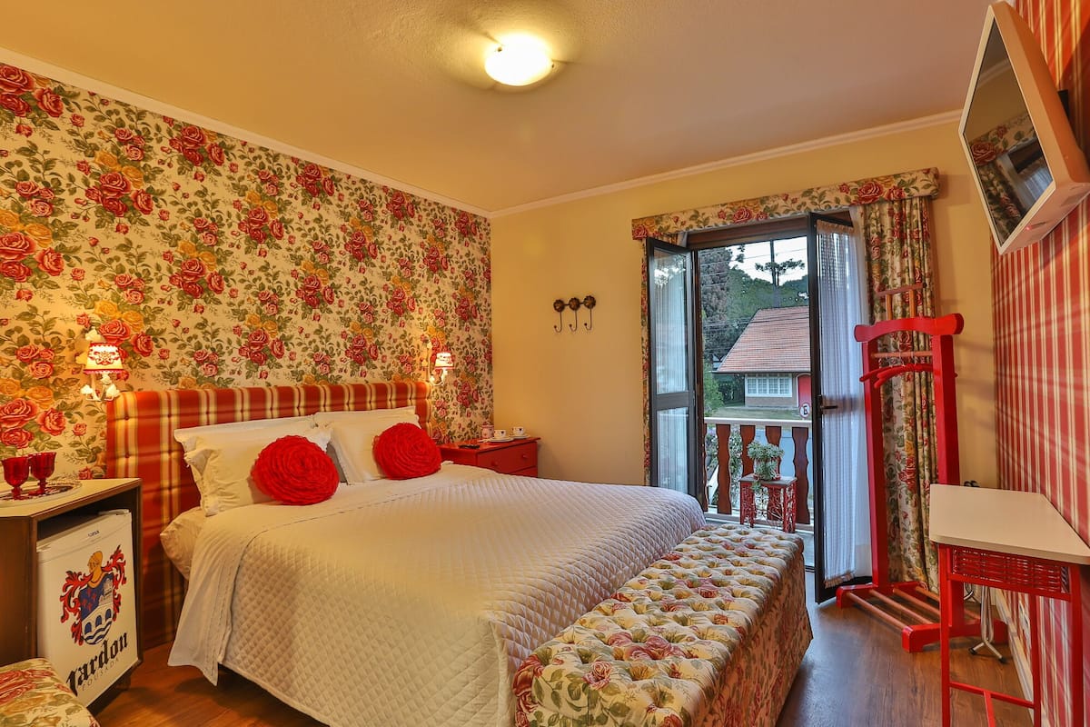 Une chambre à coucher avec un lit, une table de chevet, une commode, un fauteuil rouge et une fenêtre donnant sur une maison.