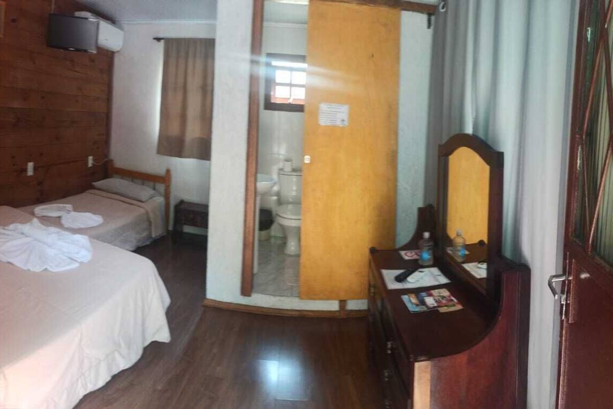 A room with wooden walls, a bed with white linens, a wooden desk with a mirror, and a door leading to a bathroom.