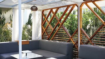 Un espacio de estar al aire libre cubierto, con bancos y mesas acolchados, rodeado de una estructura de celosía de madera y follaje.