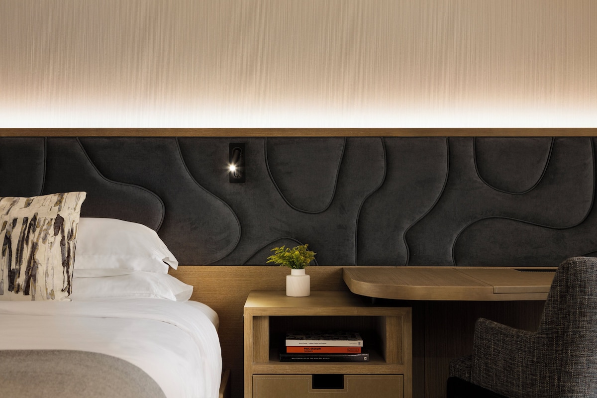 A neatly made bed with a pillow, a bedside table with a plant and books, and a chair.