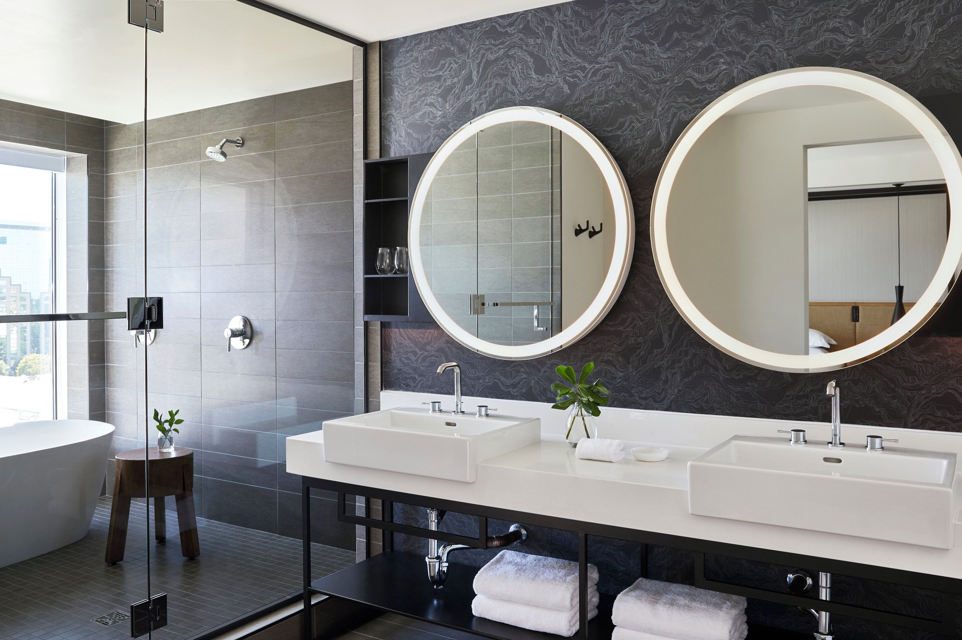 A modern bathroom with two sinks, large mirrors, a glass shower enclosure, and a small wooden stool.