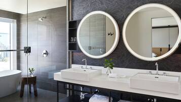 A modern bathroom with two sinks, large mirrors, a glass shower enclosure, and a small wooden stool.