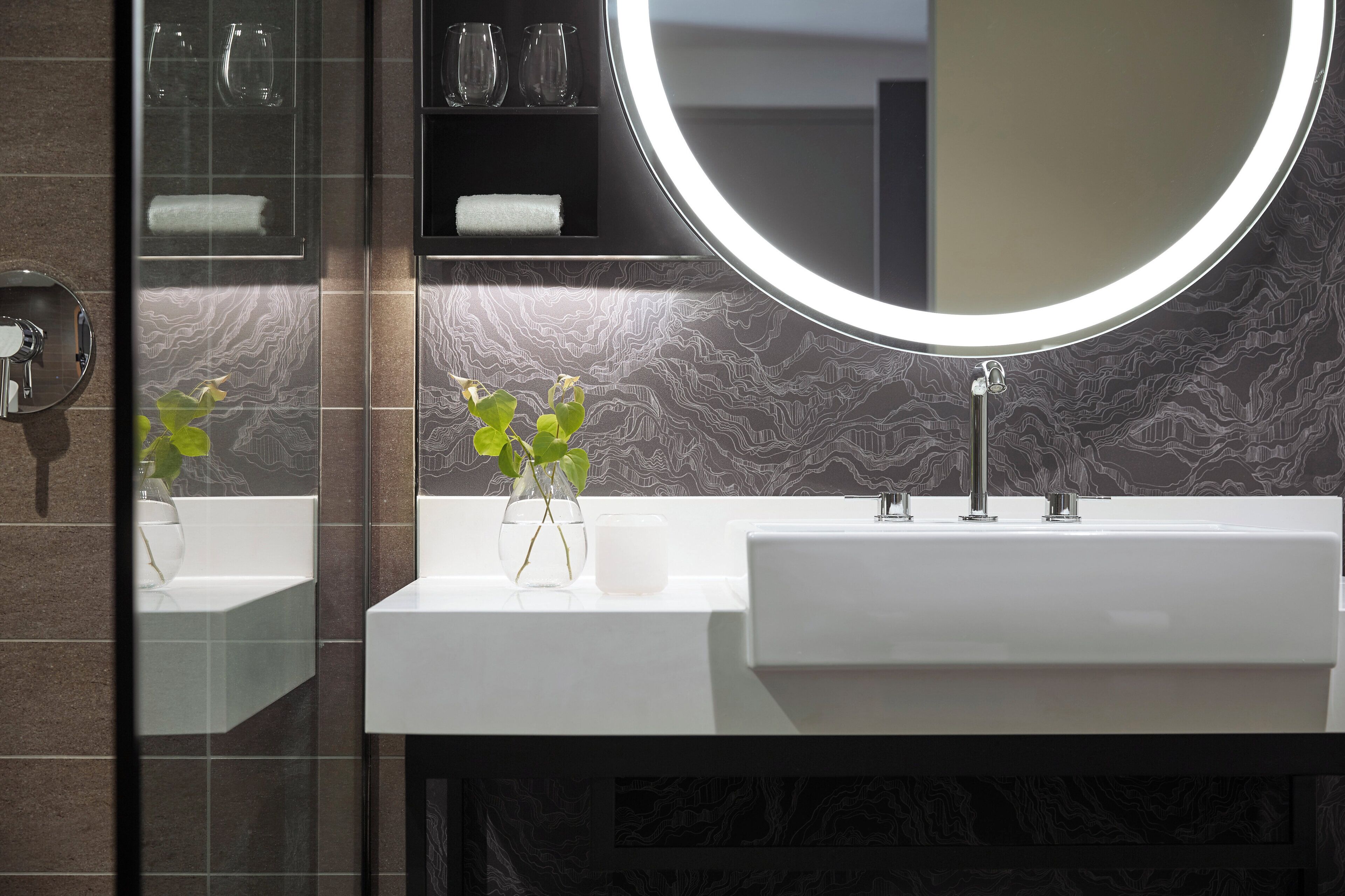 Un baño moderno con lavamanos doble, un espejo redondo y un florero con plantas verdes.