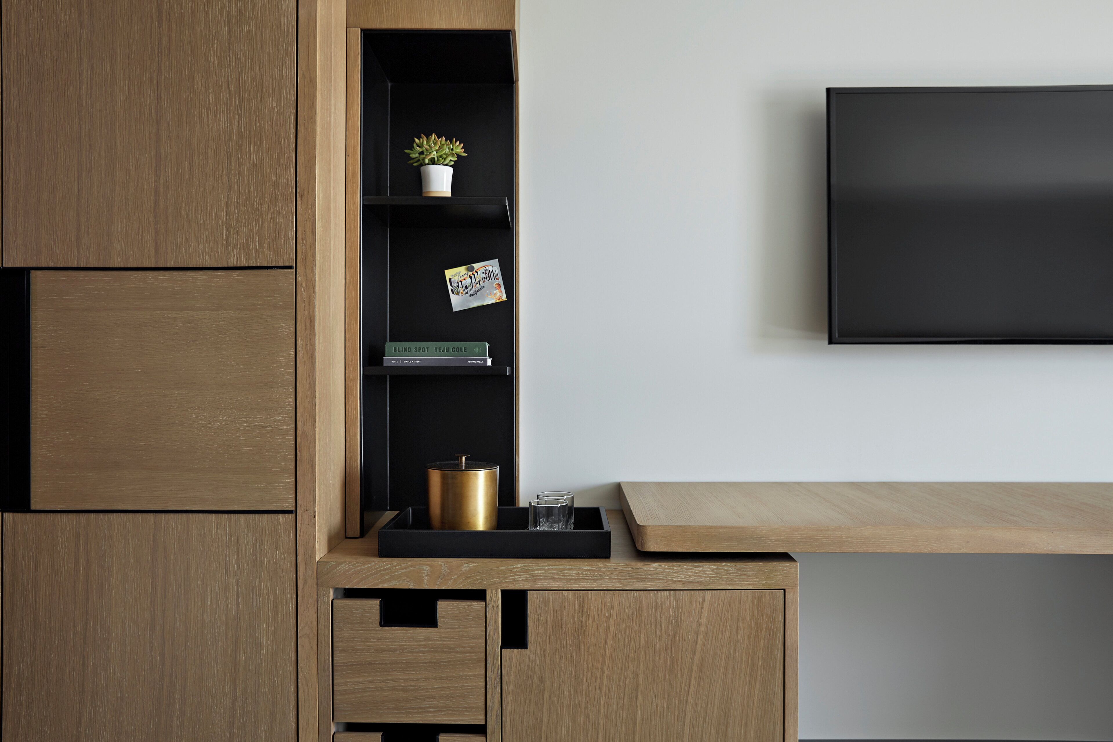 A modern, minimalist room with a wooden desk, a black TV mounted on the wall, and a shelf with a plant and books.