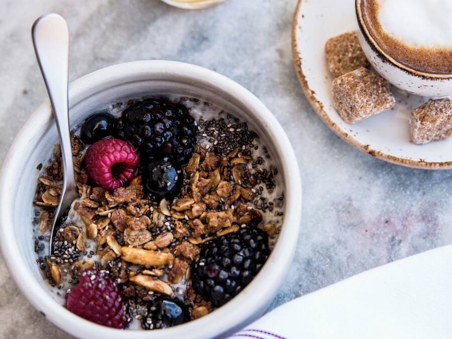 A bowl of cereal with fruits and granola, a glass of orange juice, and a cup of coffee with milk on a marble surface.