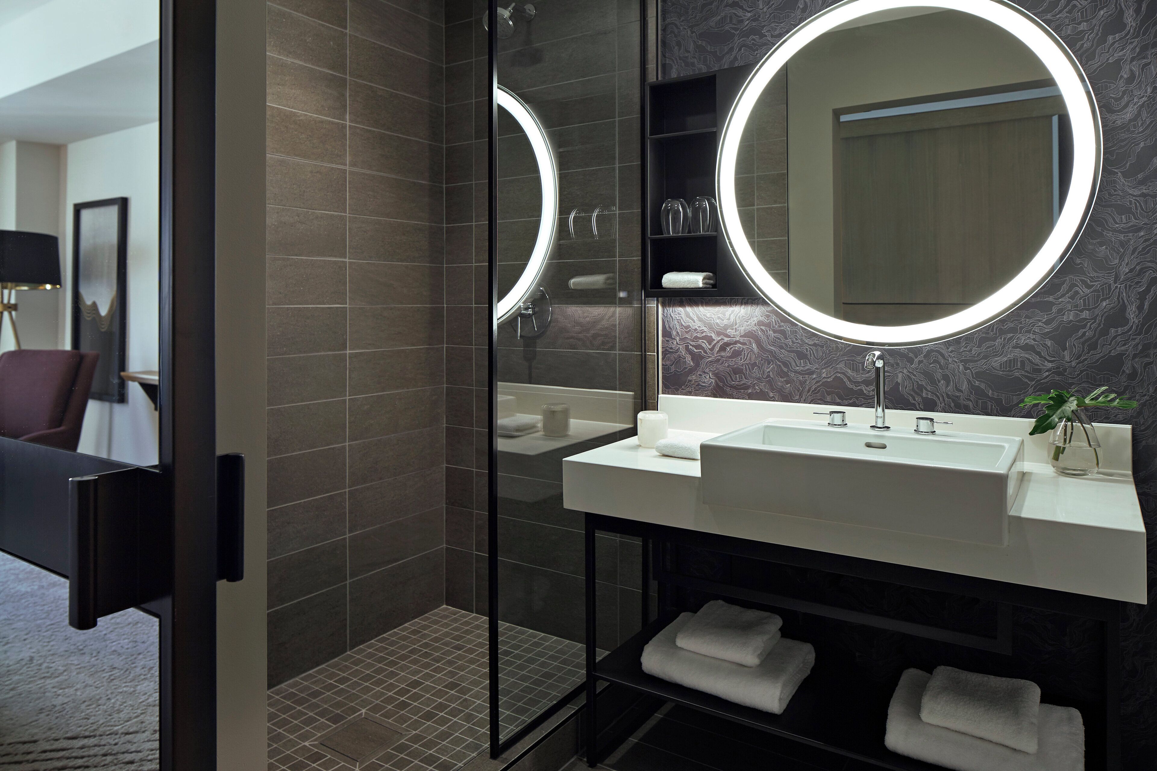 A modern bathroom with a glass shower enclosure, a large mirror with integrated lighting, and a white sink with a plant.