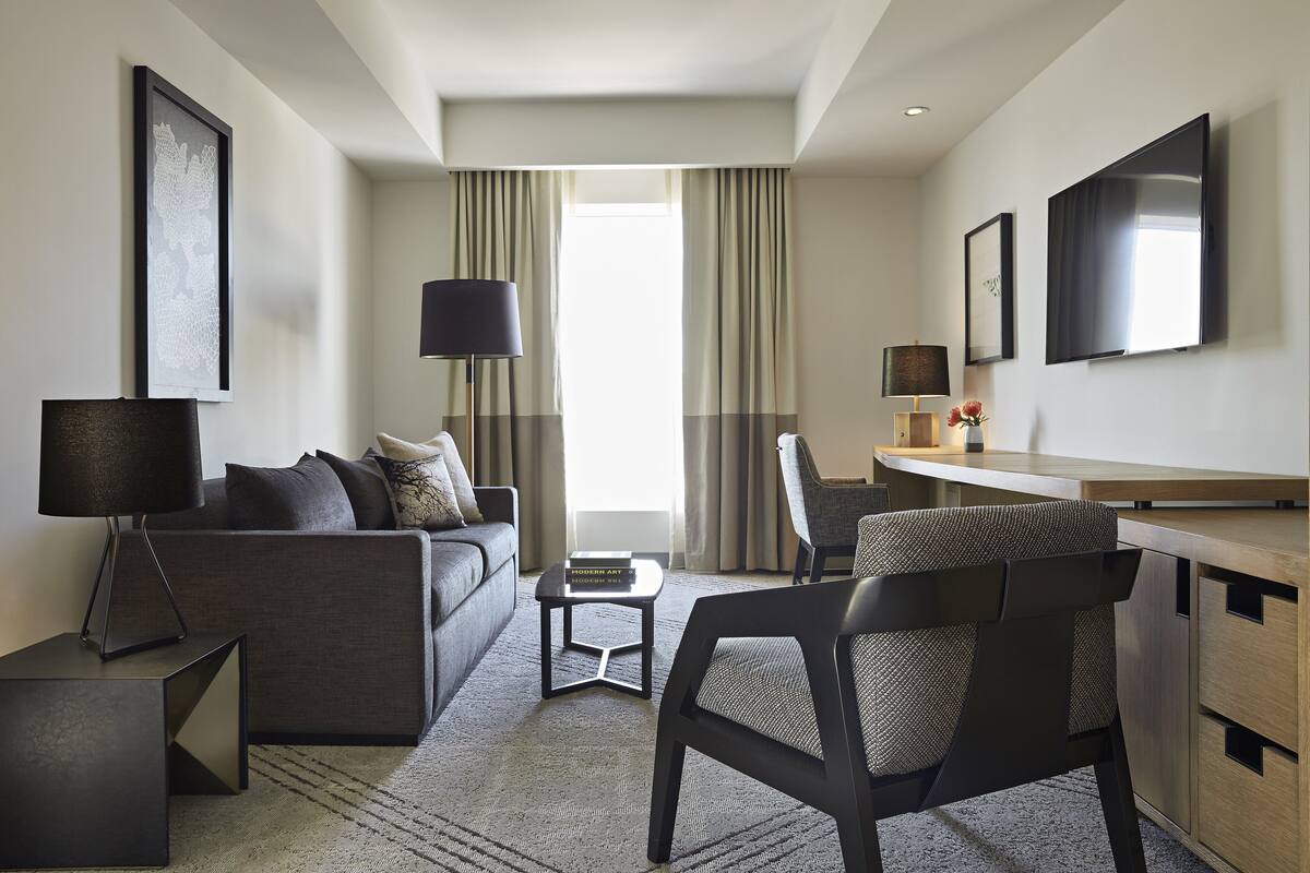 Une chambre d’hôtel moderne avec un canapé gris, un fauteuil noir, une petite table, un bureau et un téléviseur fixé au mur.