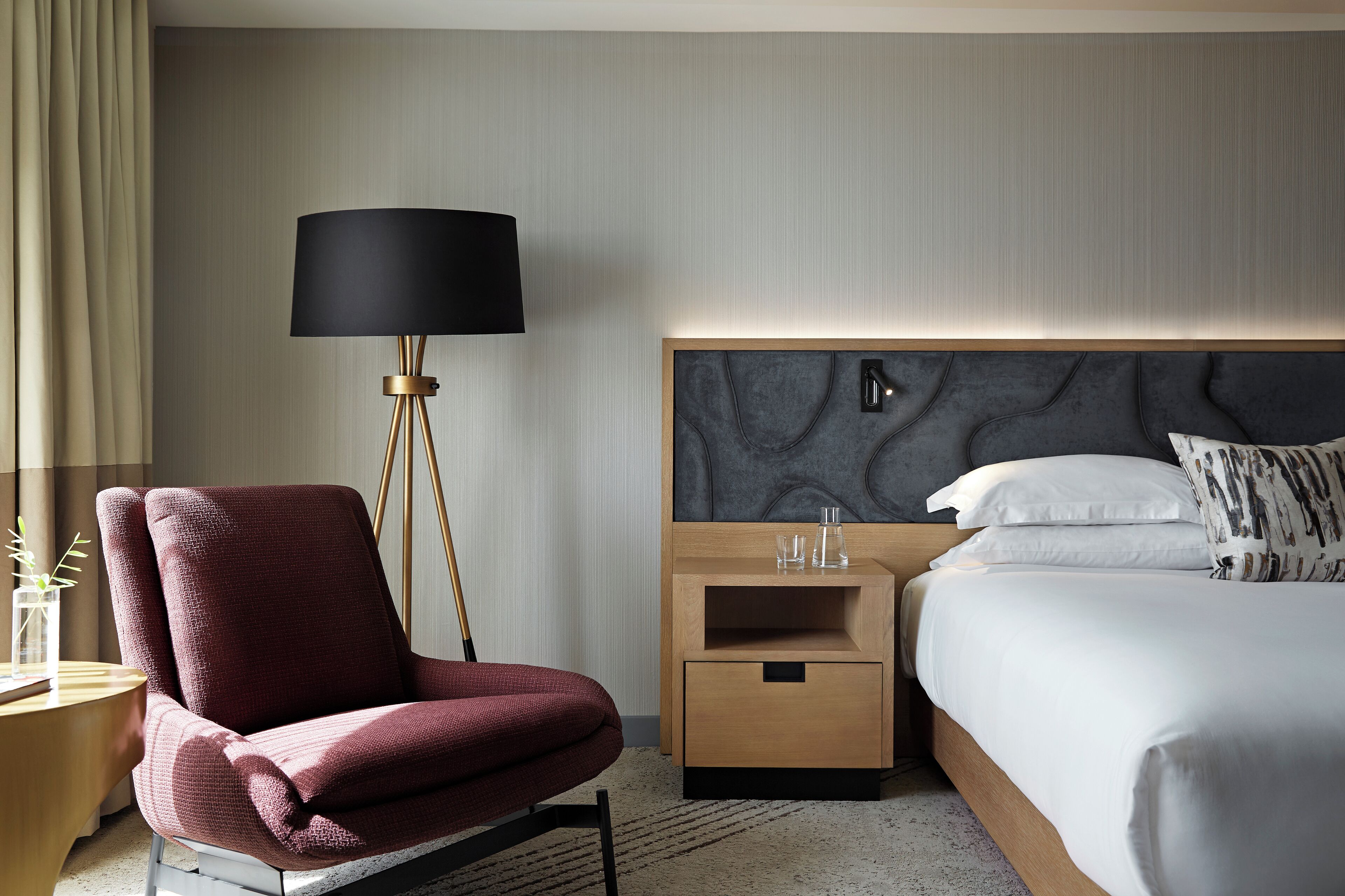 Quarto de hotel moderno com uma cama grande, uma poltrona vermelha, uma mesa de cabeceira com abajur e painel de cabeceira fixado na parede.