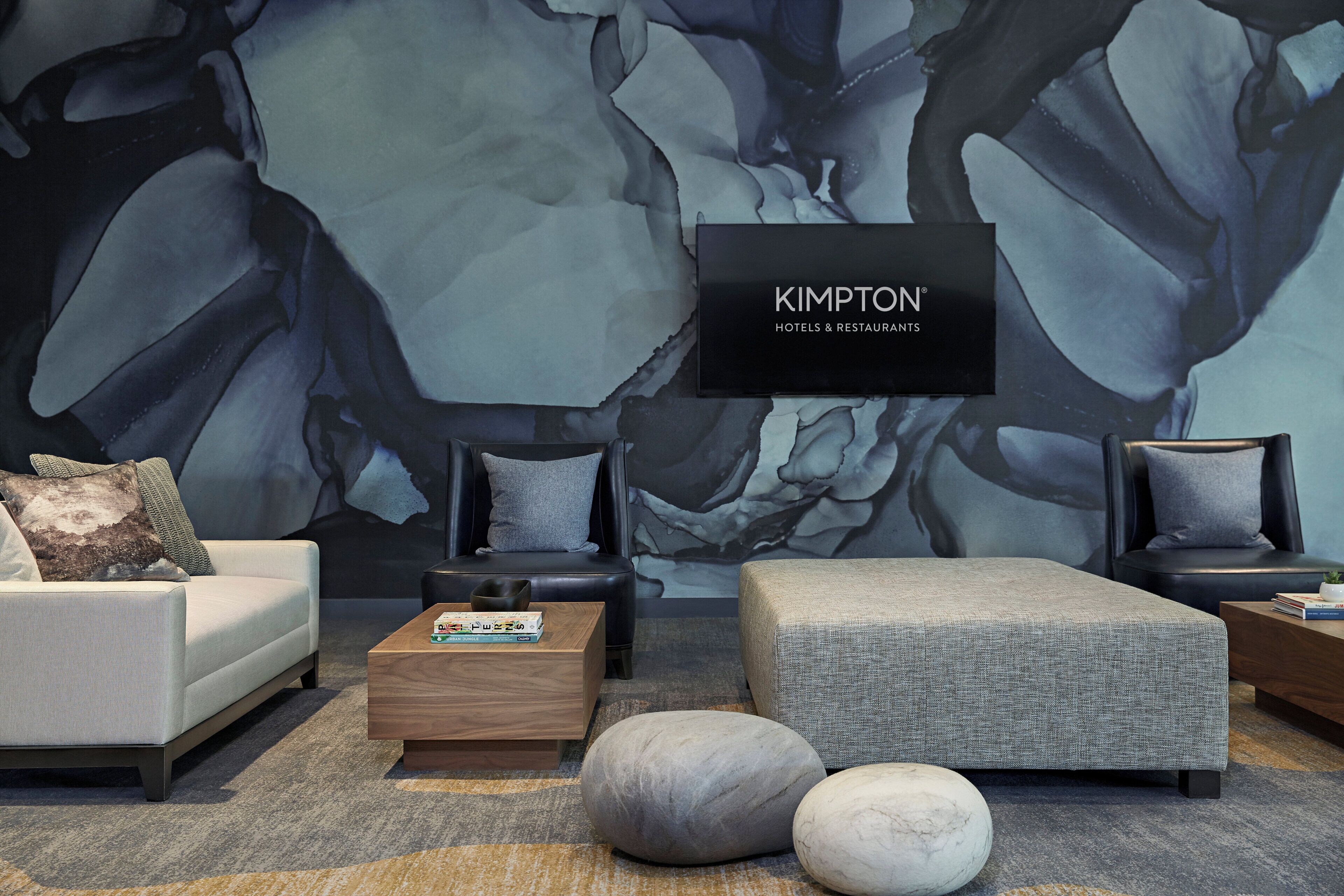 Ein modernes Wohnzimmer mit einer großen abstrakten Wandmalerei, einem Flachbildfernseher, der 'KIMPTON HOTELS & RESTAURANTS' zeigt, und zeitgemäßen Möbeln.