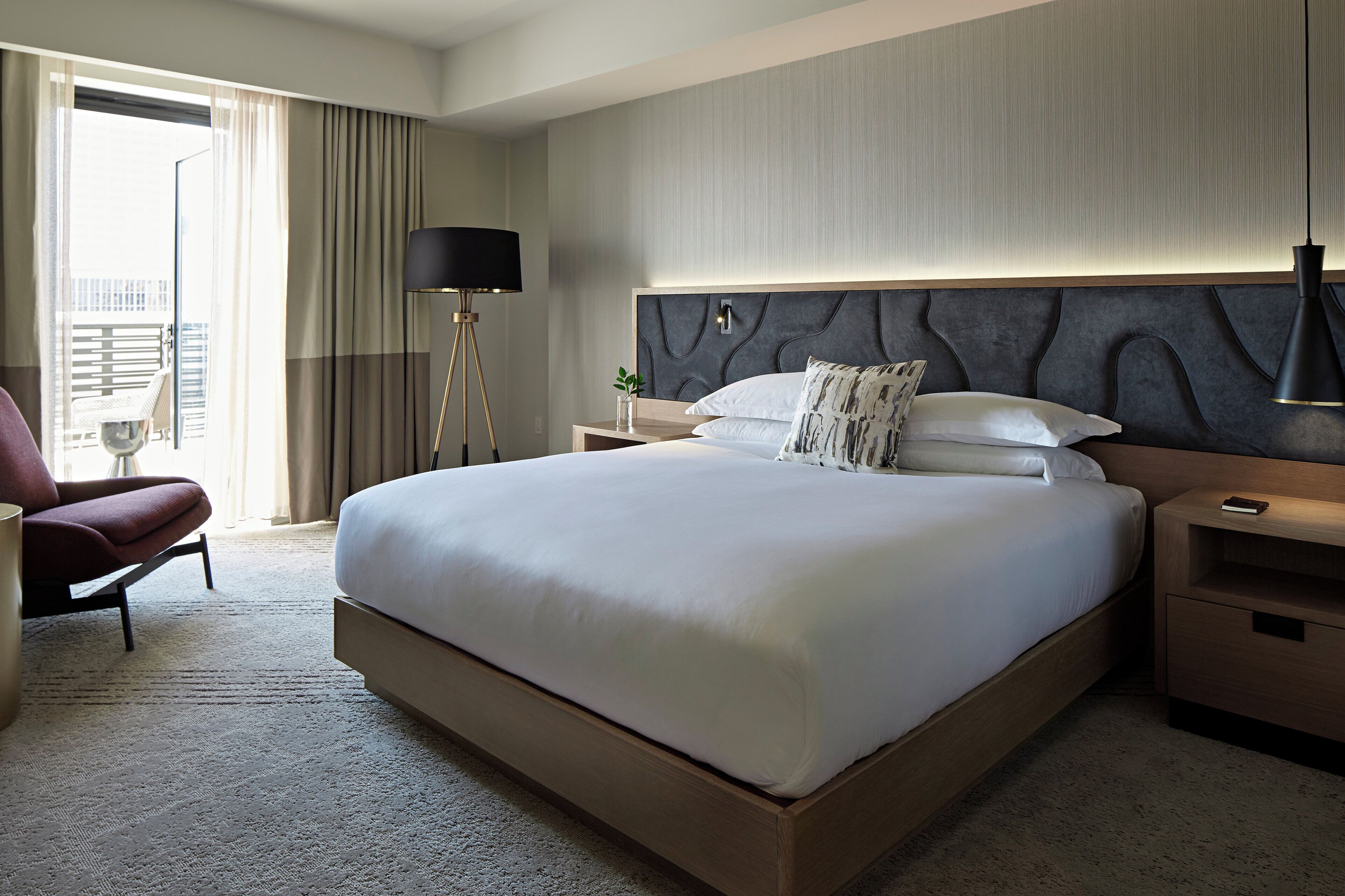 A hotel room with a large bed, a chair, a bedside table, a lamp, and a view of the outdoors.