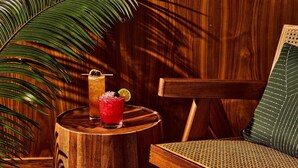 A wooden table with a drink and a lime wedge, a woven chair, and a large palm leaf. - Kimpton Sawyer Hotel by IHG (Sacramento)