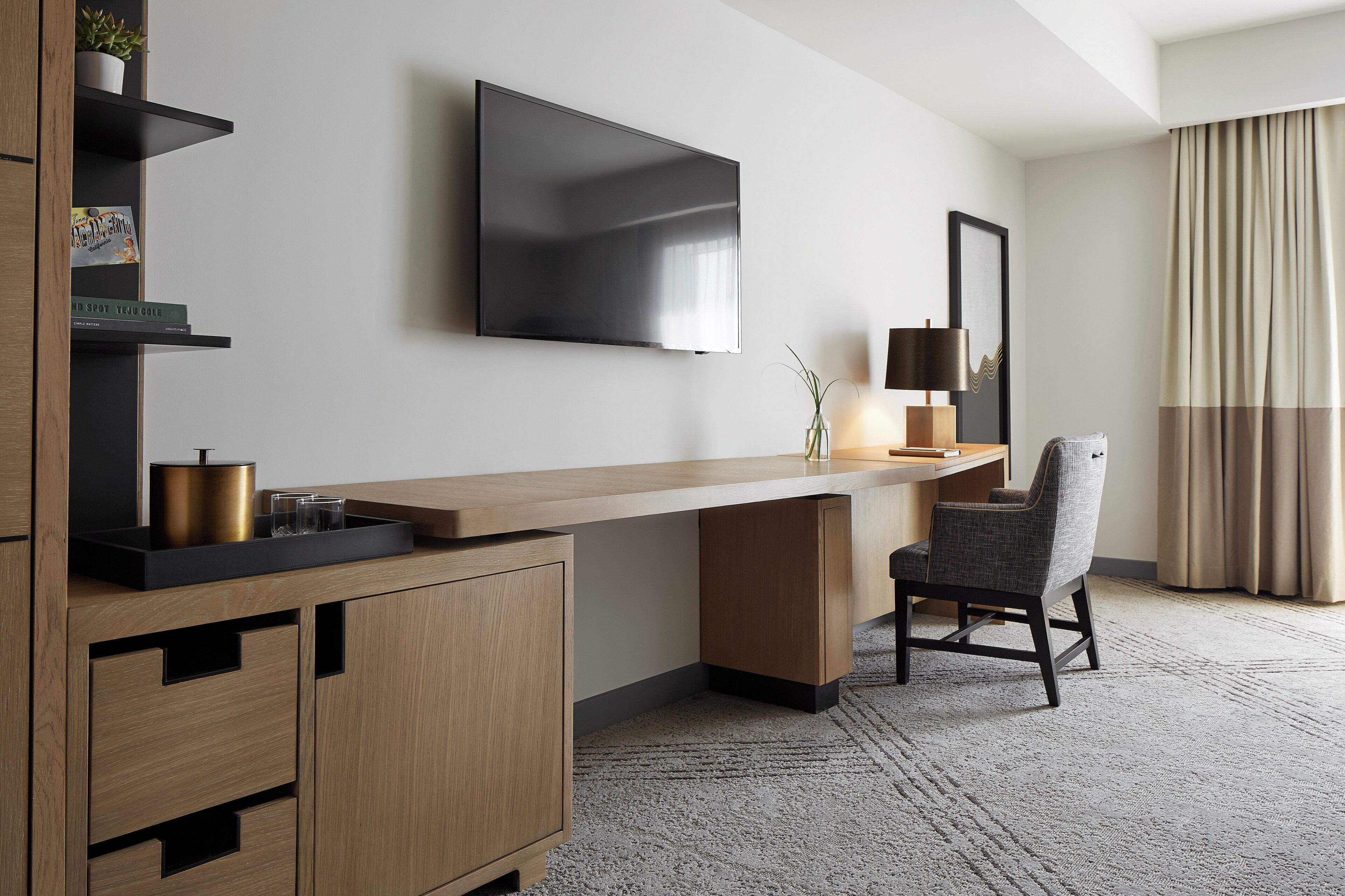 A modern hotel room with a flat-screen TV mounted on the wall, a wooden desk, a chair, a lamp, and a small plant on a shelf.