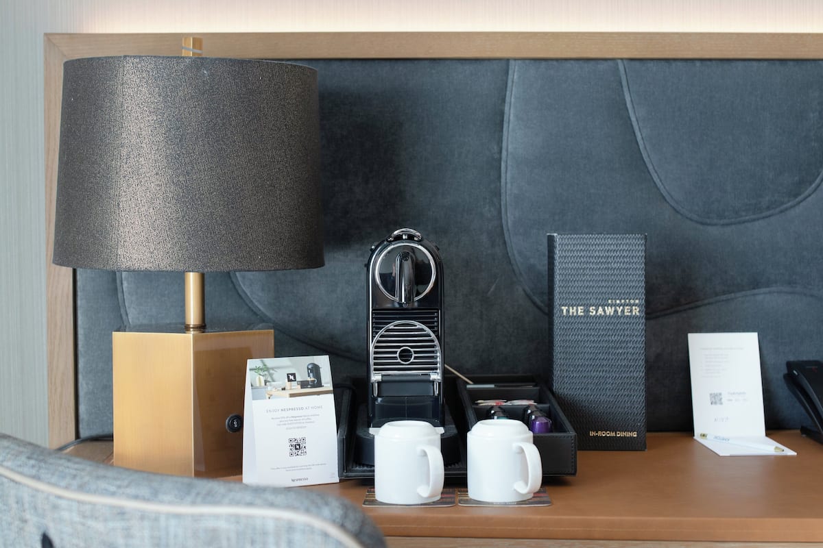 Une chambre d’hôtel bien rangée, avec une machine à café moderne, une boîte étiquetée « The Sawyer » et une lampe sur une table de chevet.