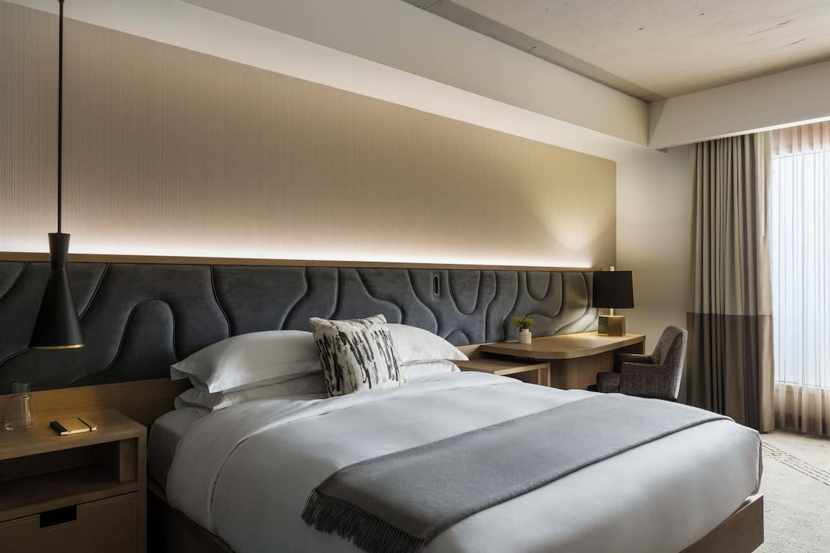 Une chambre d’hôtel moderne avec un grand lit, une tête de lit au design original, une table de chevet avec une lampe, un fauteuil et une vue sur l’extérieur par une fenêtre.
