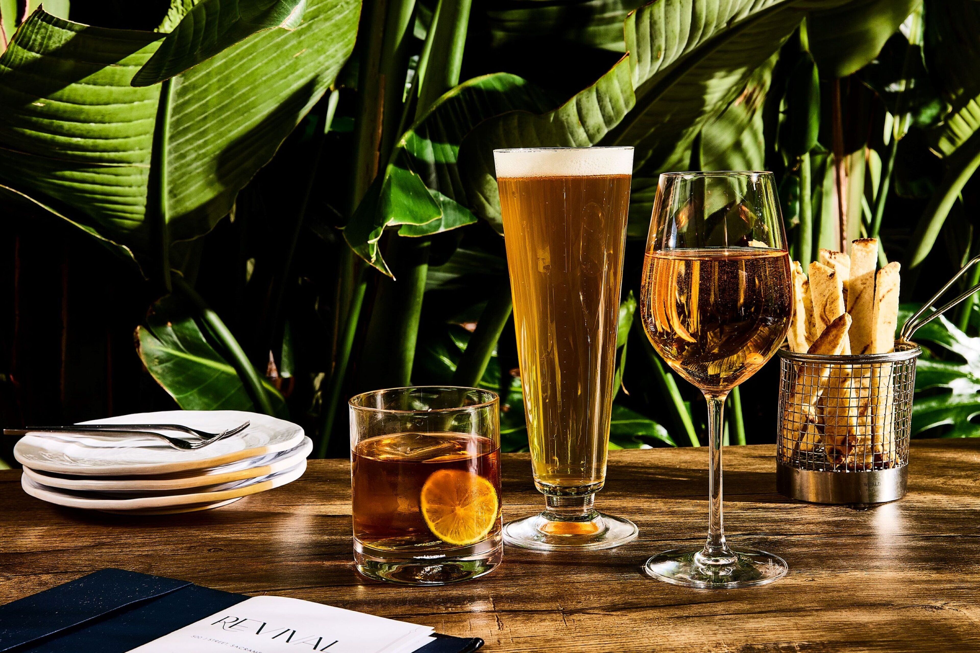 A table with a tall glass of beer, a short glass with a slice of orange, and a tall glass of champagne. There are plates and a basket of breadsticks.