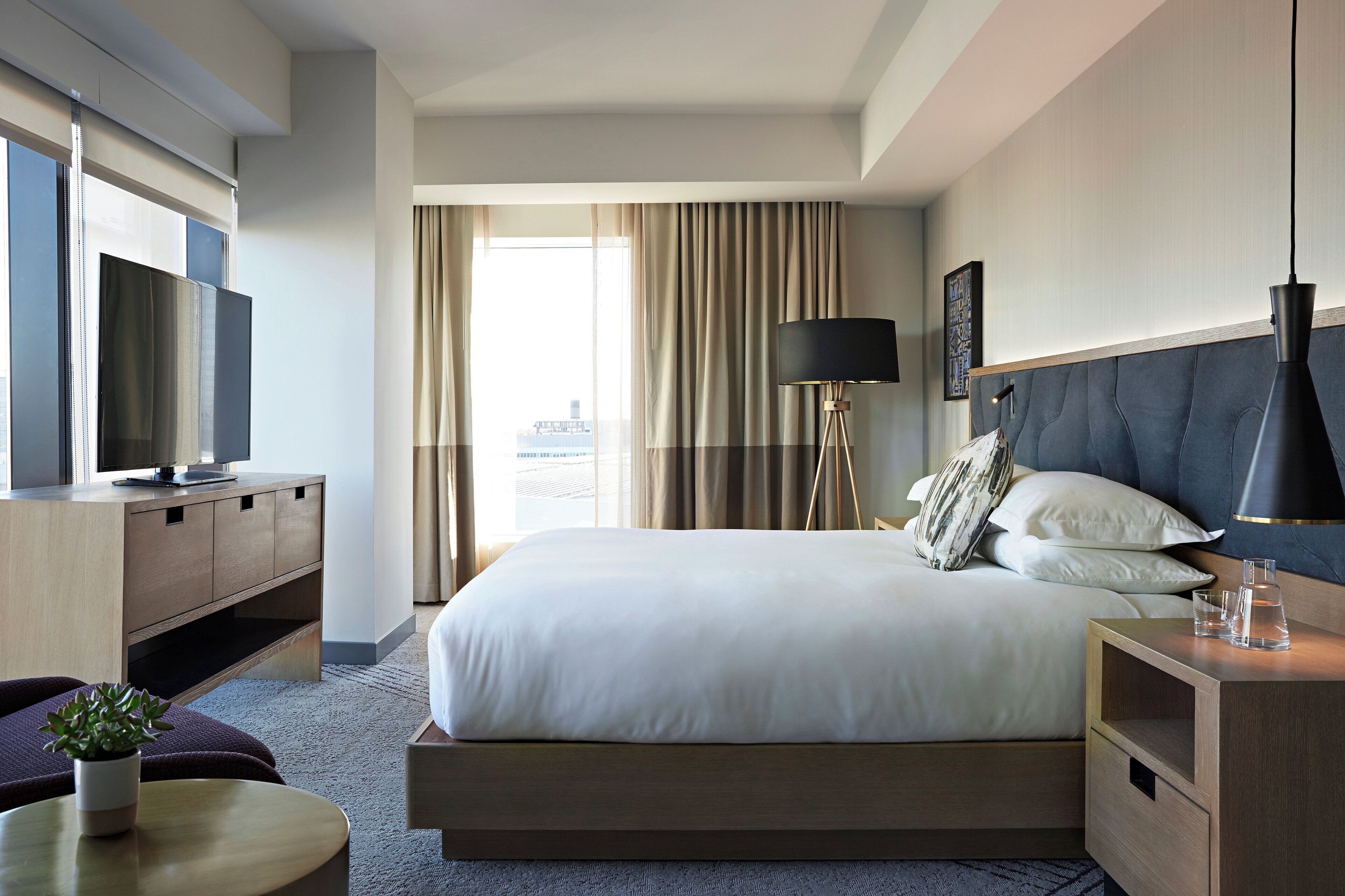 A hotel room with a bed, a TV, a lamp, a bedside table, and a view of the city.