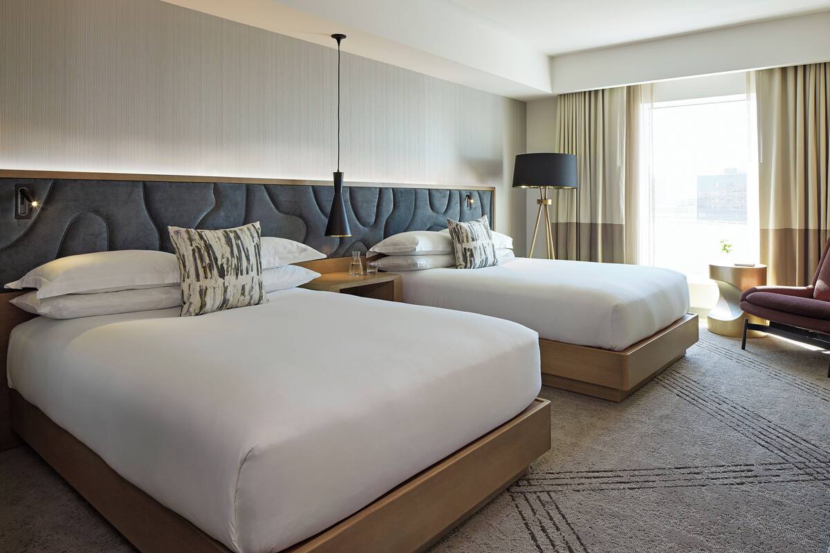 Une chambre d’hôtel avec deux lits, une tête de lit moderne, une table de chevet, une lampe, une chaise et une vue sur la ville.