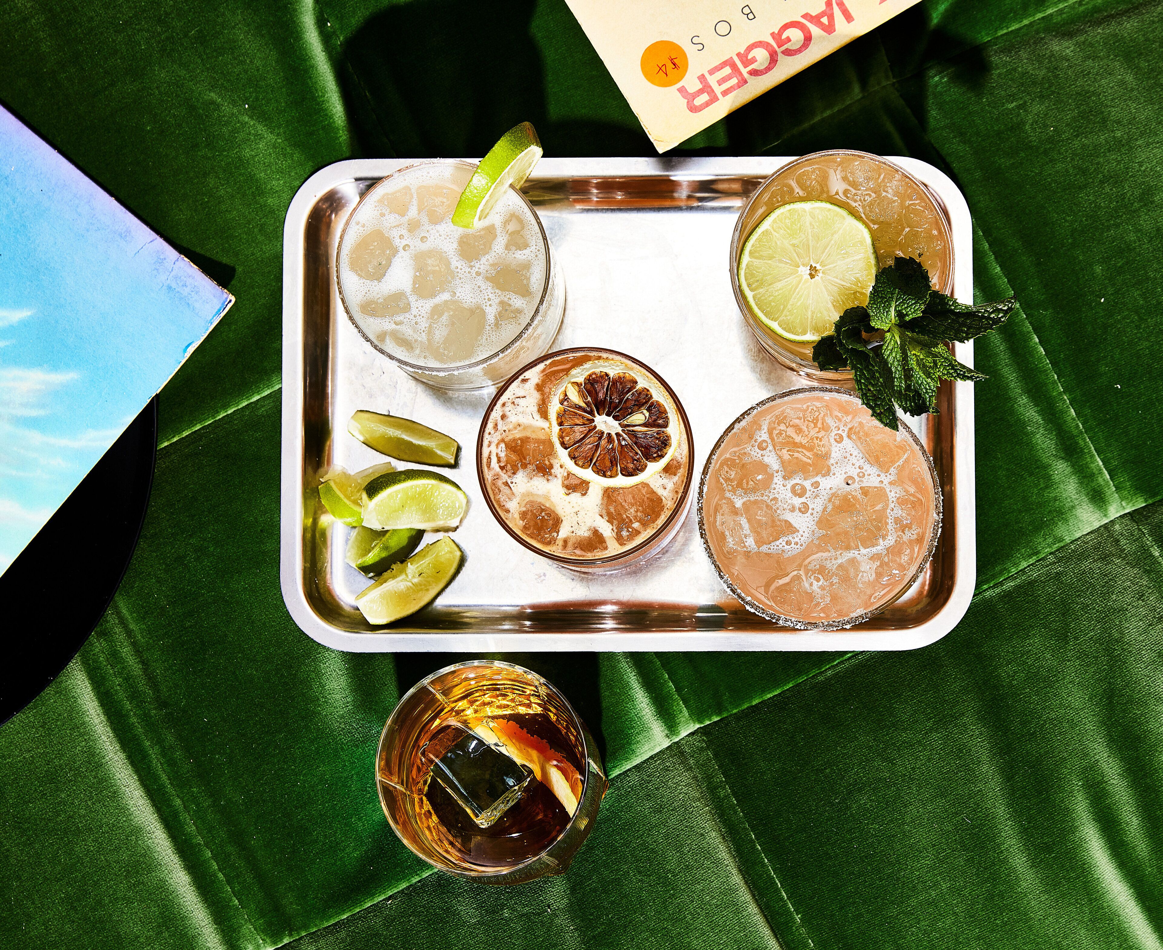 Una bandeja con cuatro bebidas, cada una adornada con una rodaja de lima, y un vaso de whisky con un cubito de hielo.